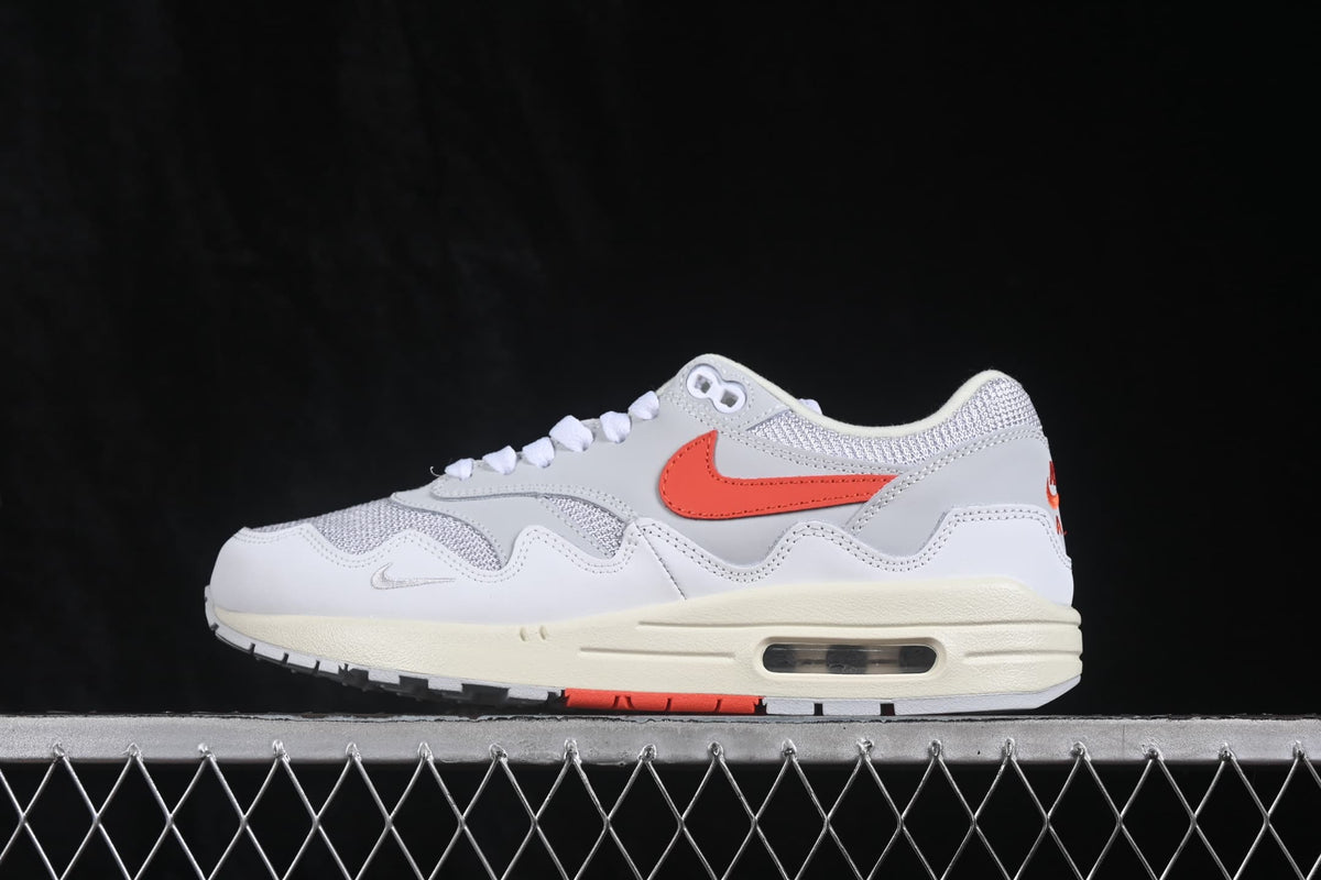 Side profile of a white and grey Nike Air Max 1 sneaker with an orange Swoosh, cream midsole, and visible air unit.