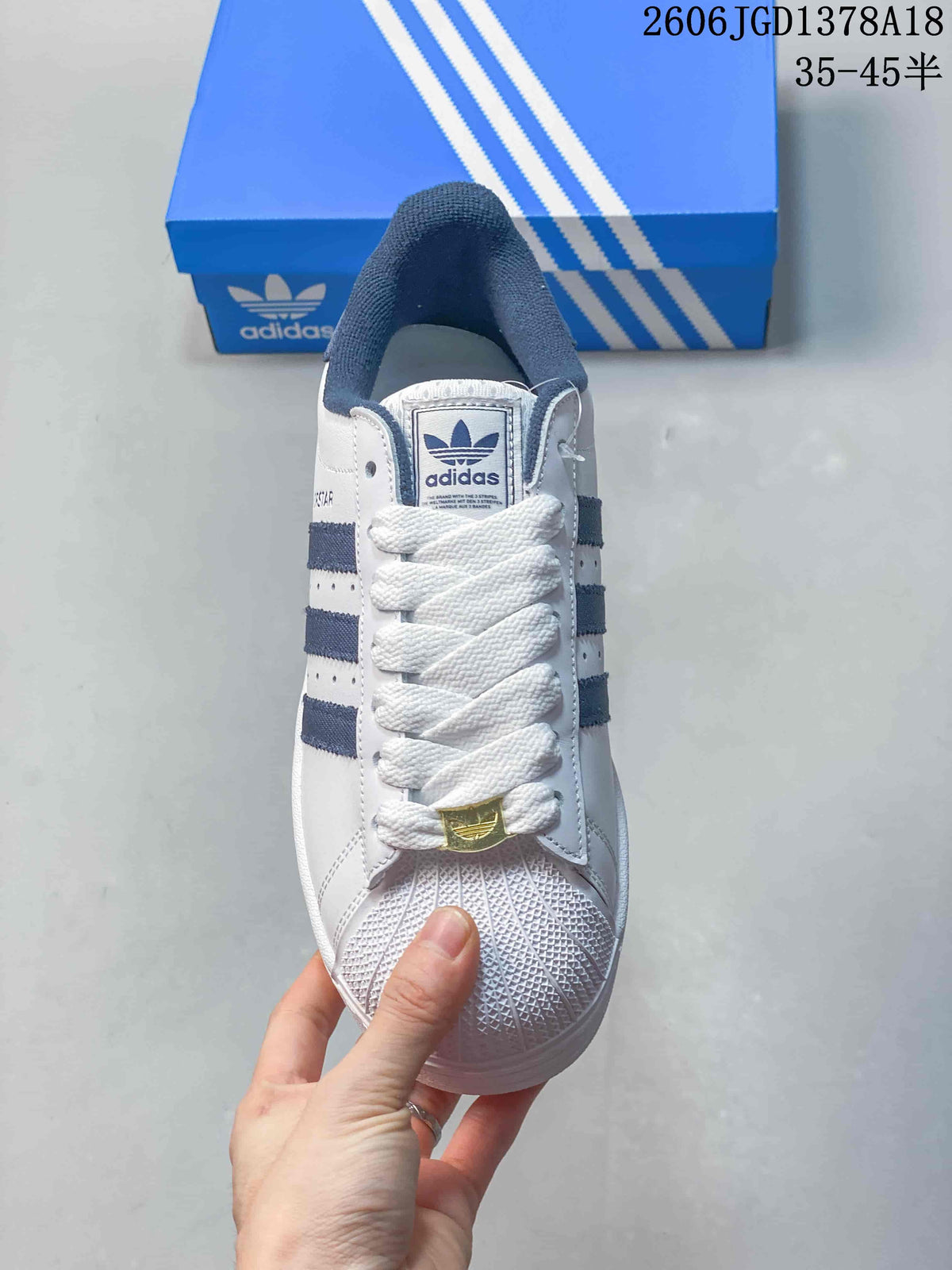 Front view of a white Adidas Superstar sneaker with navy details and textured shell-style toe.