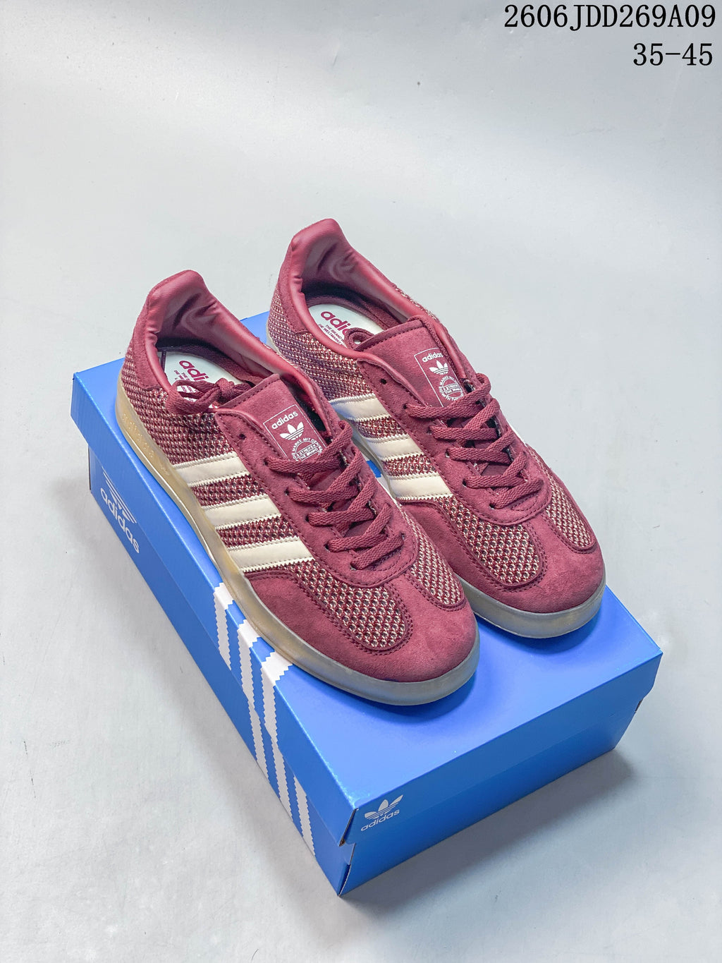 Burgundy adidas Gazelle Indoor sneakers with light stripes and a gum-colored outsole, pictured on a blue shoe box