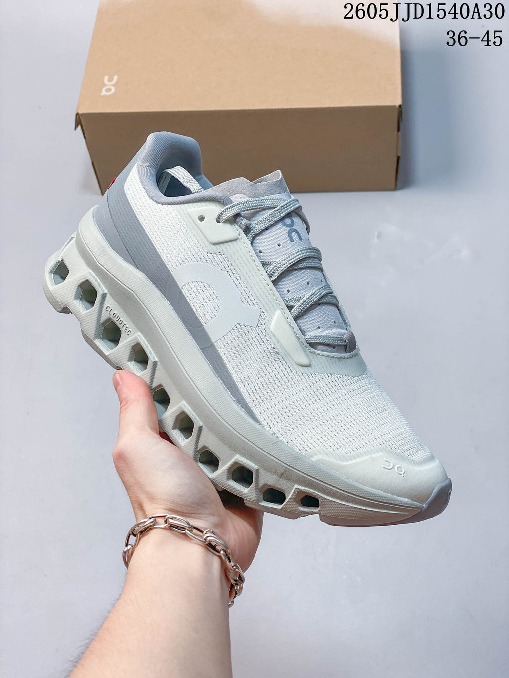 Side view of a pale gray and white On Cloudmonster 2 shoe showing its open-channel sole and blue-gray accents.