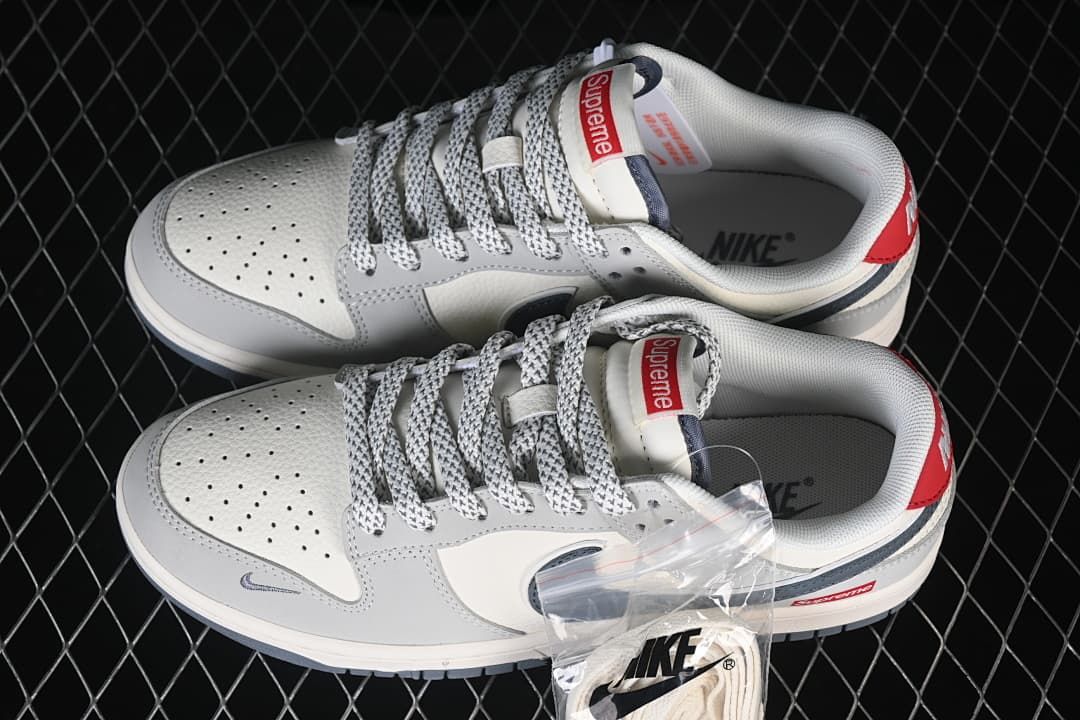 Pair of grey and off-white Nike SB Dunk Low sneakers with patterned laces and red Supreme tongue details.