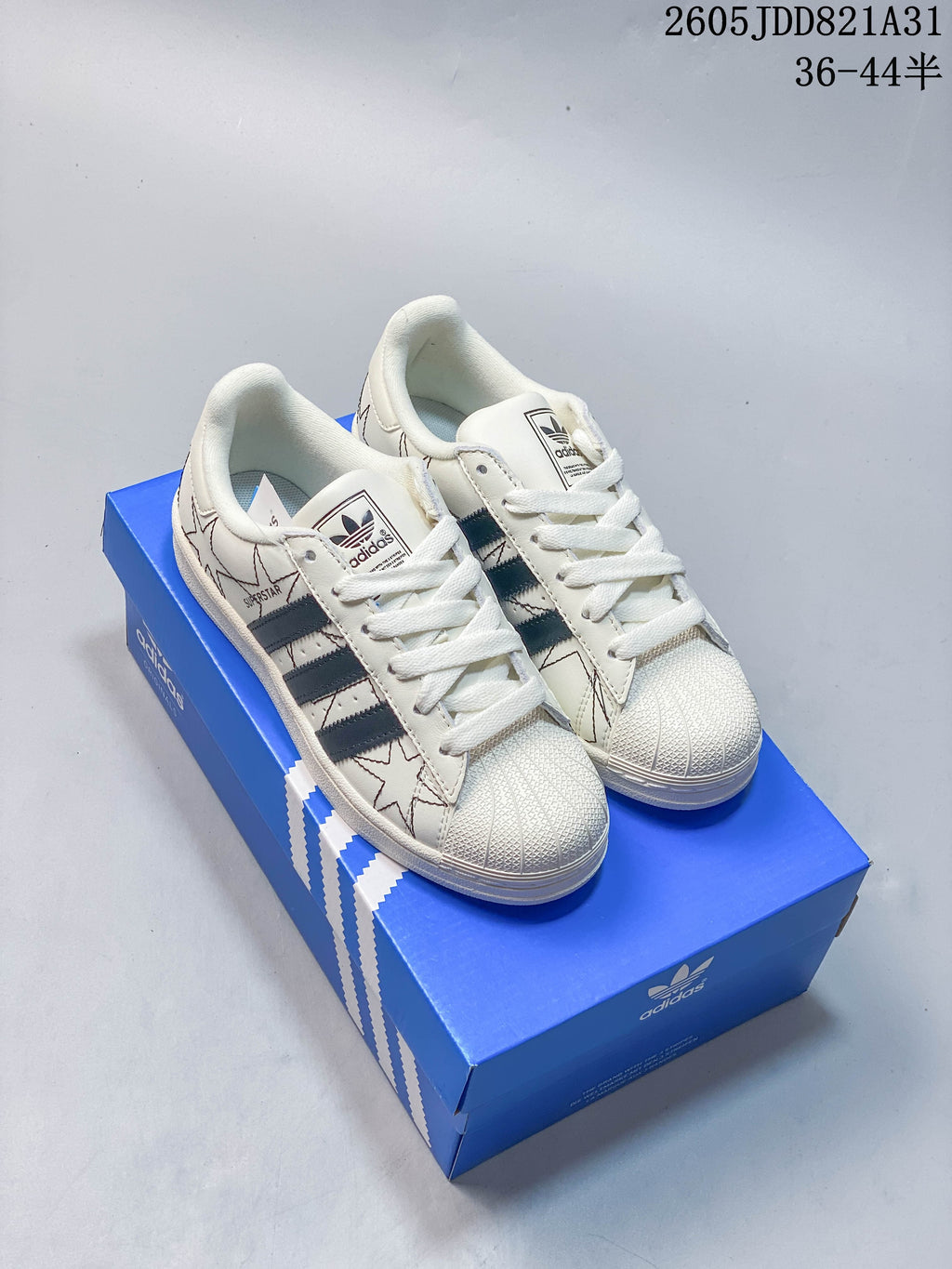 White Adidas Superstar sneakers with dark stripes and outlined star graphics, shown on a blue shoe box.