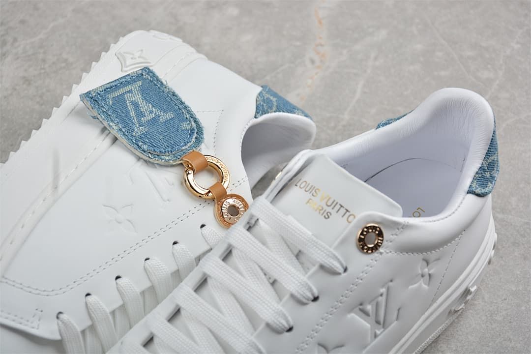 Close-up of white sneakers showing blue denim-look accents, embossed motifs and gold-tone hardware