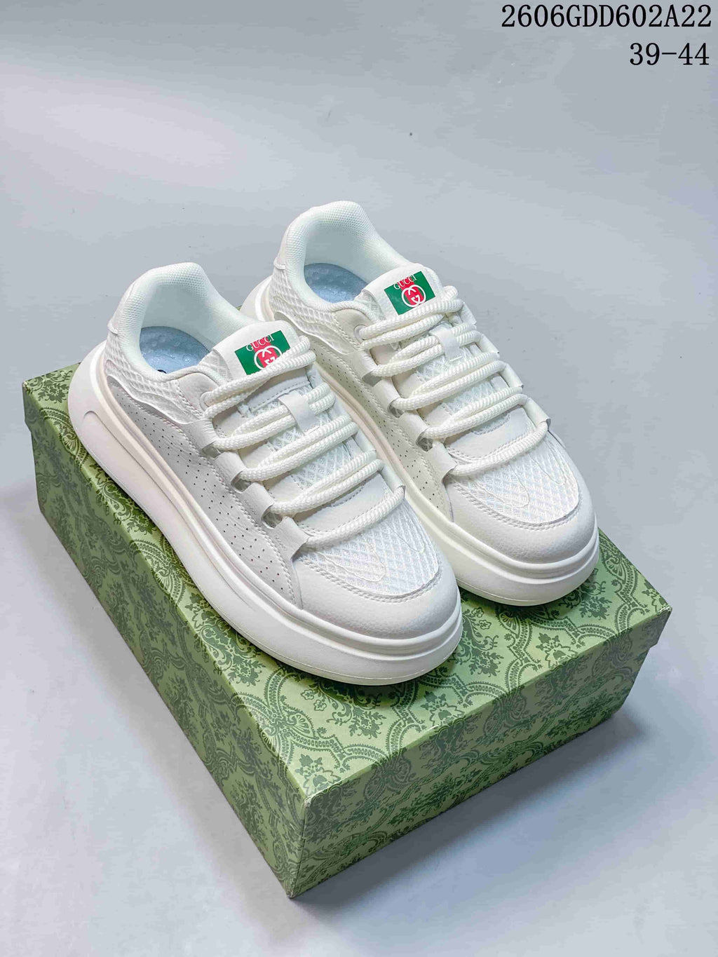 Pair of white low-top sneakers with white laces and patterned green box.