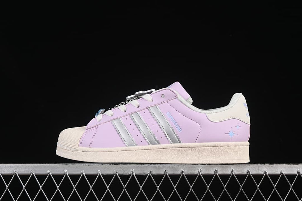 Side view of lilac-pink adidas Superstar sneakers with silver stripes, an off-white shell toe, and lace charms