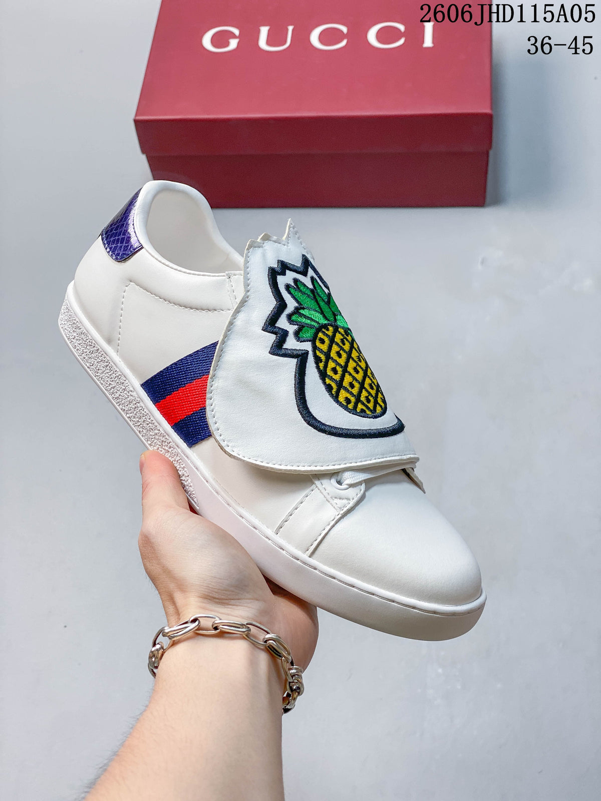 White low-top sneaker with a pineapple embroidered tongue flap, red-and-blue side stripe, and purple heel accent.