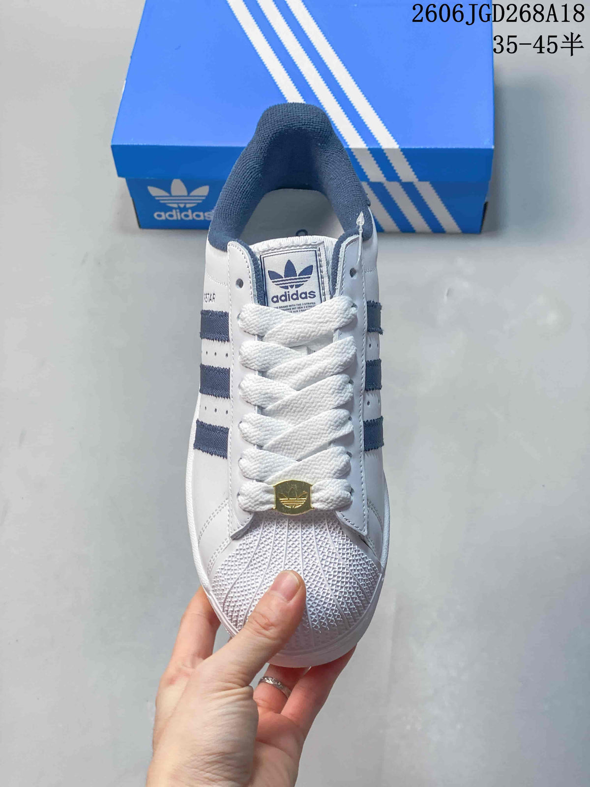 Front view of a white Adidas Superstar sneaker with textured shell toe and lace ornament.