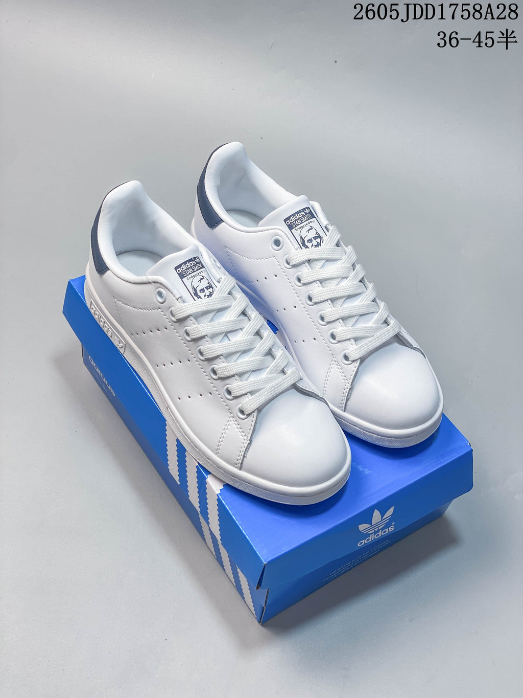 Pair of white low-top sneakers with dark blue heel tabs, photographed on a blue shoe box; image overlay shows a size range.