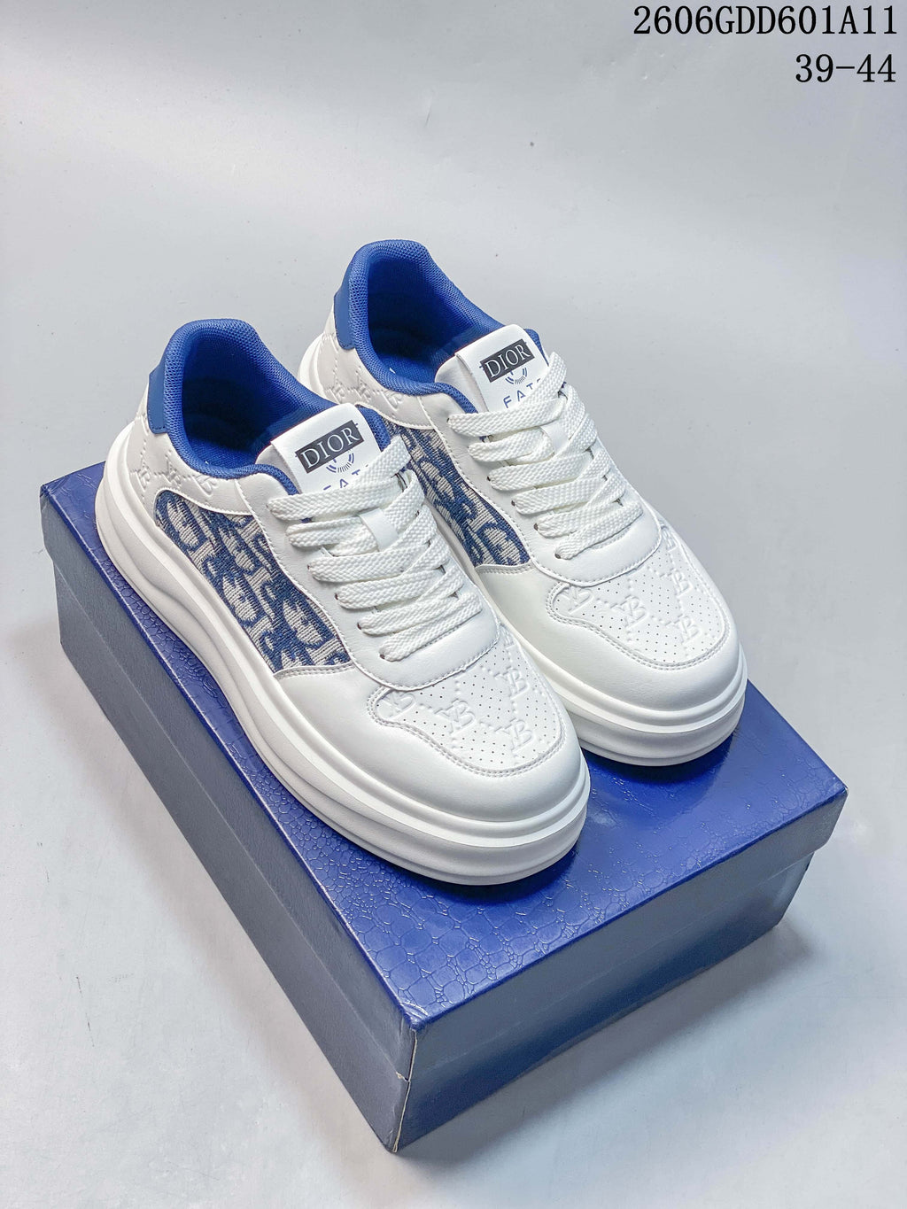 Pair of white and blue Dior-branded low-top sneakers with patterned side panels, shown on a blue box; image marking shows sizes 39–44 and style code 2606GDD601A11.