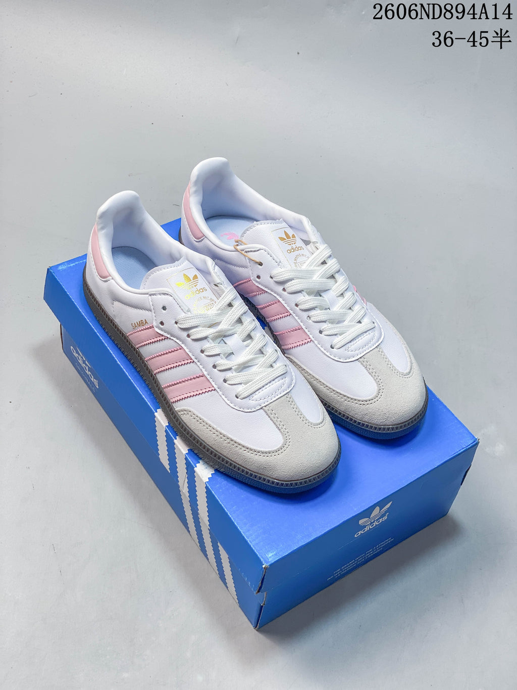 Pair of white adidas Samba OG low-top sneakers with pink stripes and heel details, light beige toe overlays, and dark gum-colored outsoles, shown on a blue shoe box.