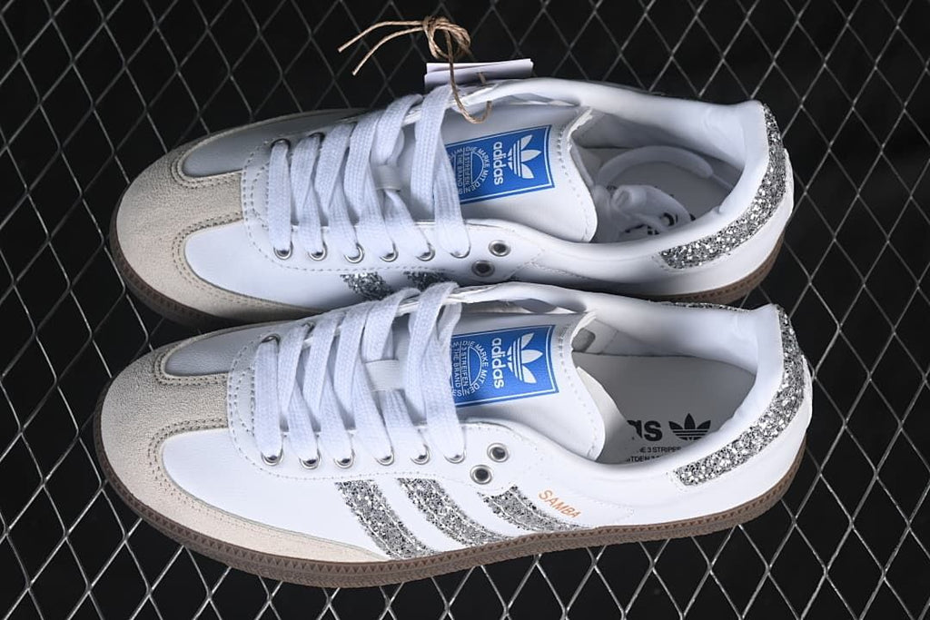 Pair of white Samba OG sneakers with silver glitter-effect accents, beige toe overlays, and gum-brown outsoles.