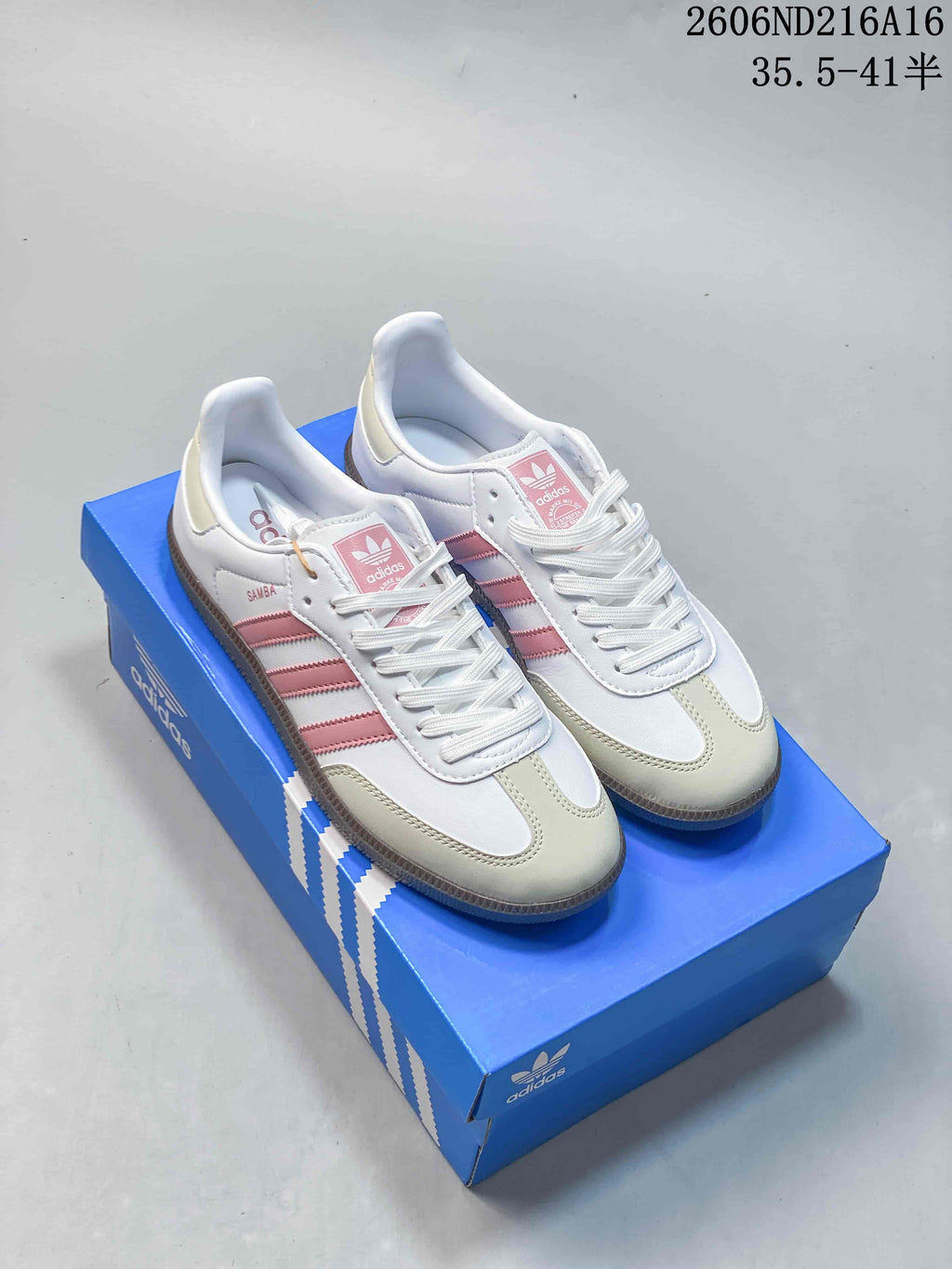 Pair of white Adidas Samba-style low-top sneakers with pink stripes and pale neutral overlays, displayed on a blue shoe box