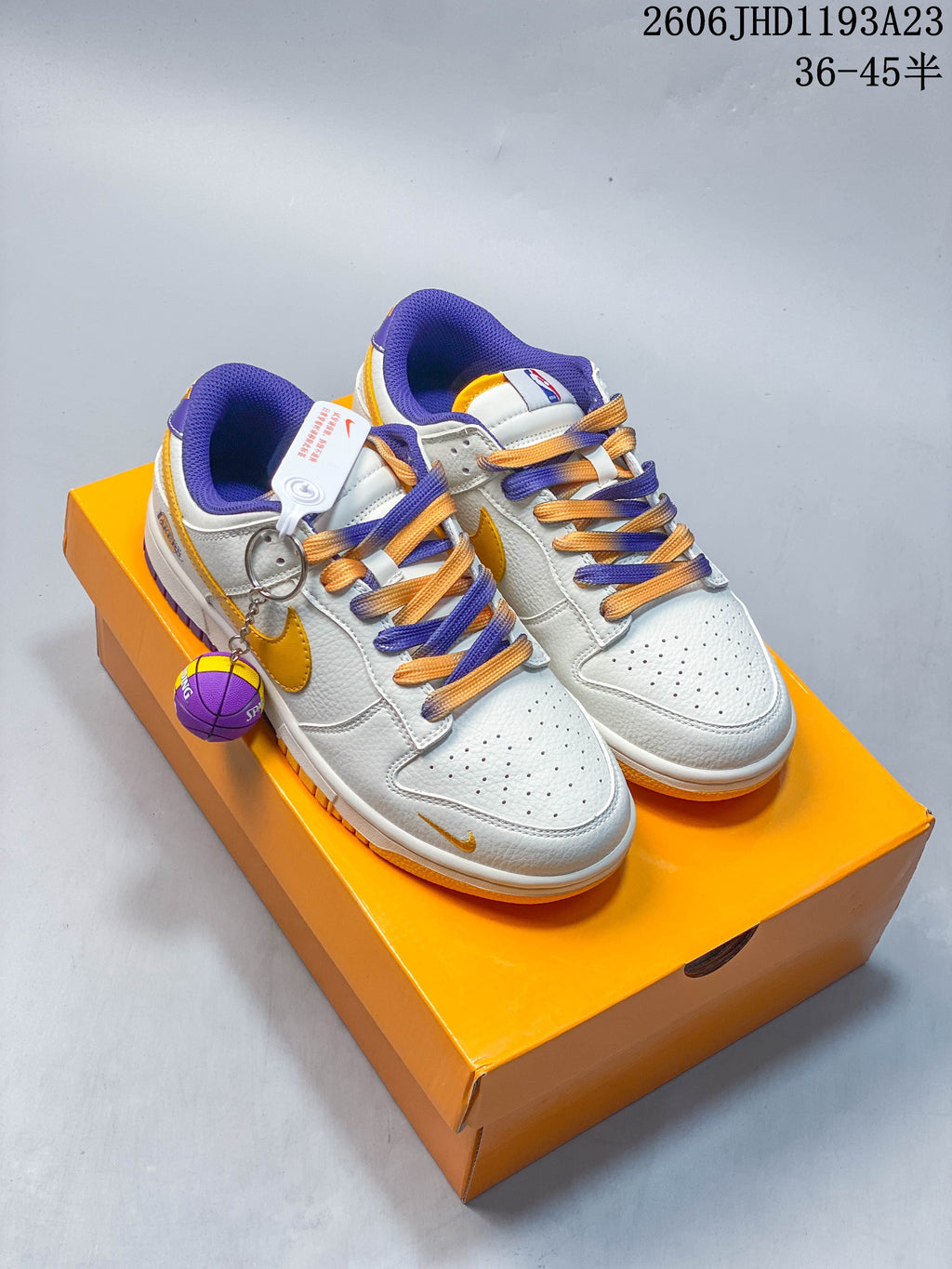 Pair of white Nike SB Dunk Low sneakers with gold and purple accents, contrasting laces, basketball charm, and orange Lakers-branded box