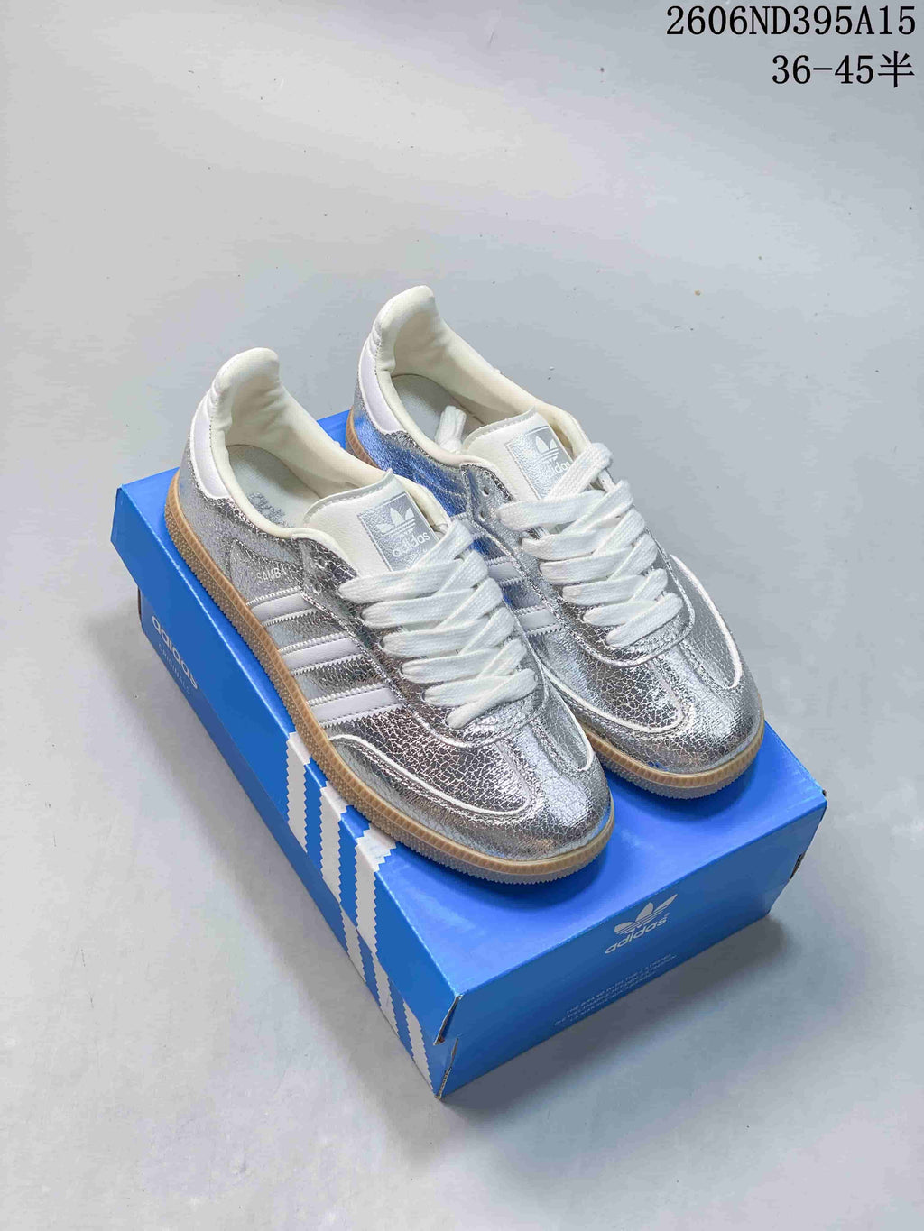 Pair of silver Adidas Originals Samba-style low-top sneakers with white stripes and gum-colored soles on a blue shoe box