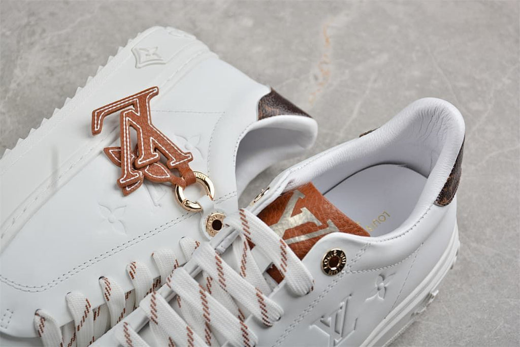 Close-up of white sneakers with brown letter charm, striped laces, and embossed details