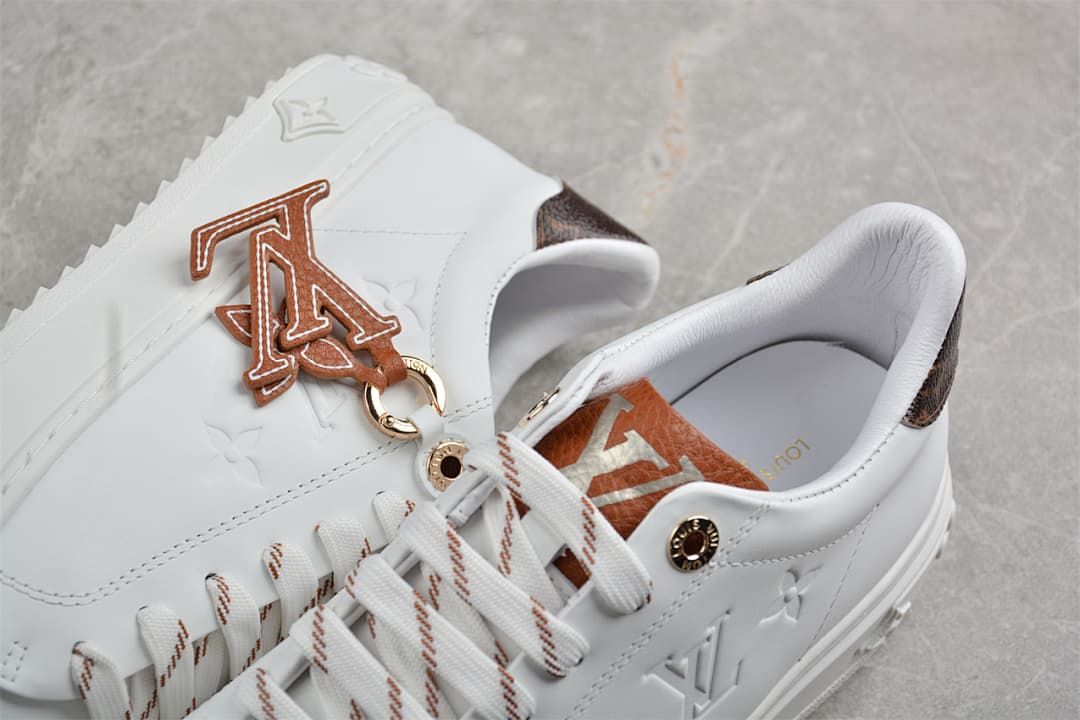 Close-up of white sneakers with brown letter charm, striped laces, and embossed details