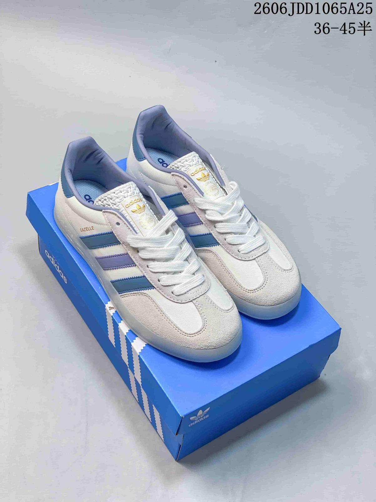 Pair of white and light-gray Adidas Gazelle low-top sneakers with blue and lavender stripes, displayed on a blue shoebox.