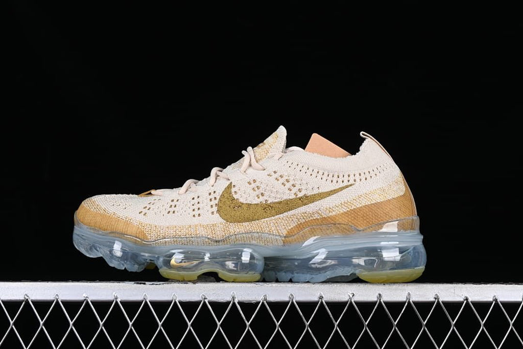 Side profile of light neutral Nike Air VaporMax 2023 Flyknit sneakers with gold-toned accents and a clear sole.