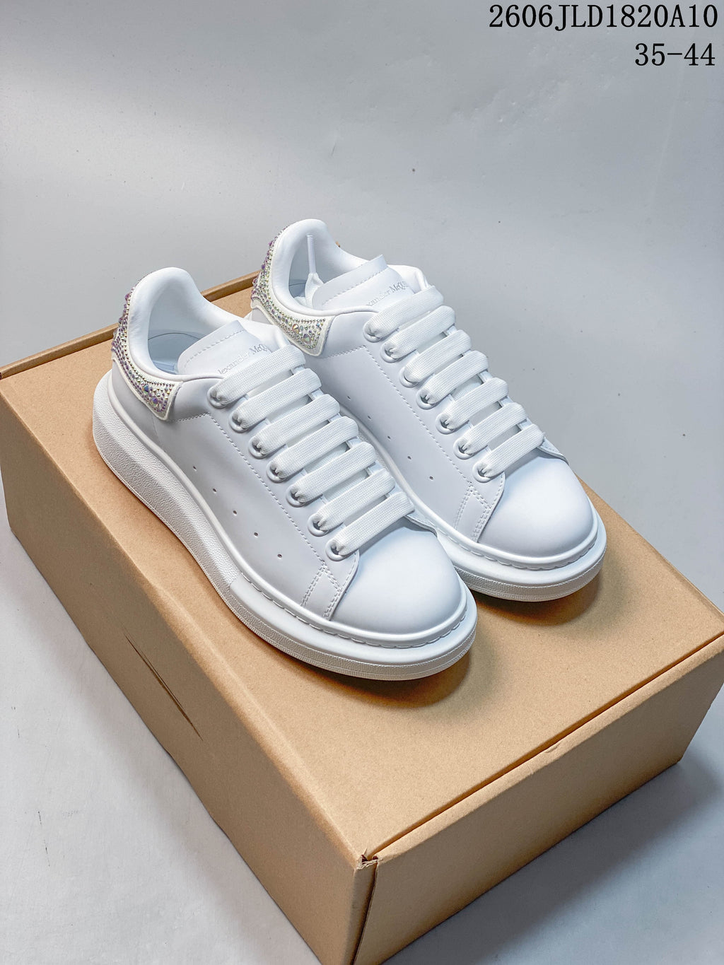 Pair of white platform sneakers with wide laces and sparkling heel details, shown on a tan box