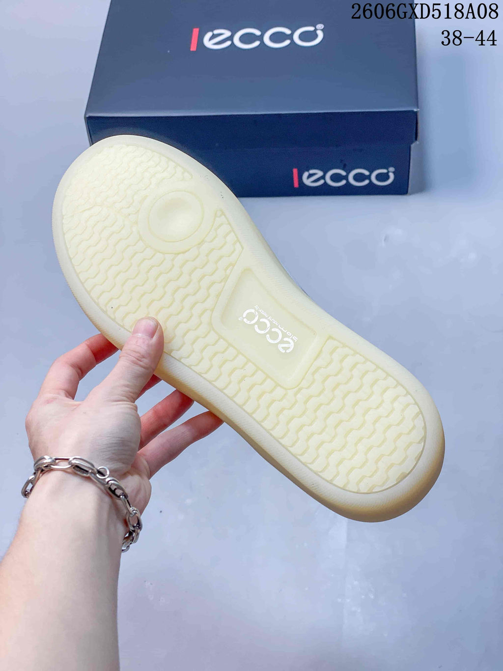 Underside of a sneaker showing a light-colored patterned tread and branded box behind it.