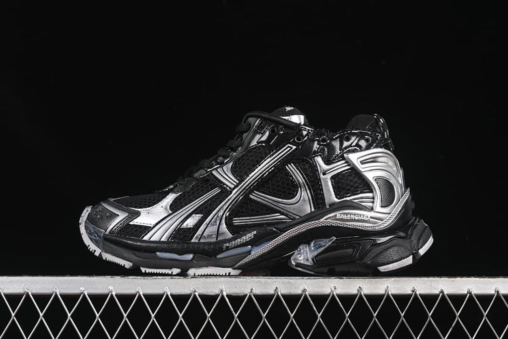 Side profile of a black and silver Balenciaga Runner-style sneaker against a dark background.