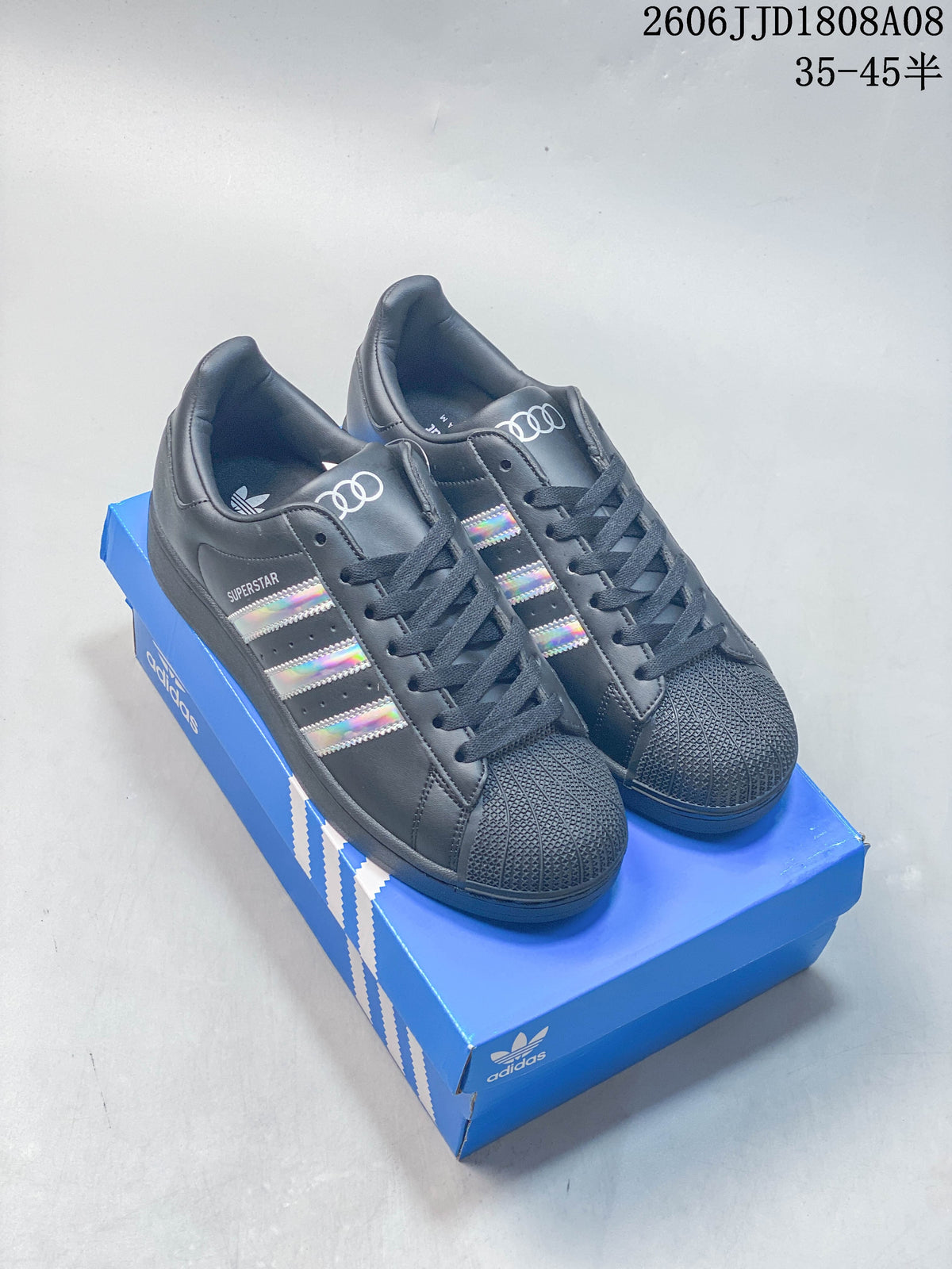 Pair of charcoal Adidas Superstar sneakers with iridescent stripes, displayed on a blue box.