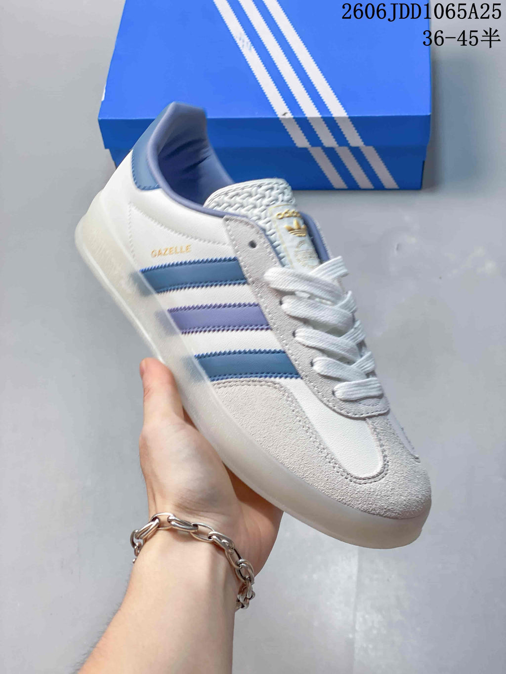 Angled view of a white and light-gray Adidas Gazelle sneaker with blue and lavender stripes.