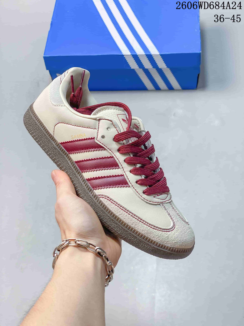 Cream Adidas Samba-style low-top sneaker with burgundy stripes and laces beside a blue shoe box.