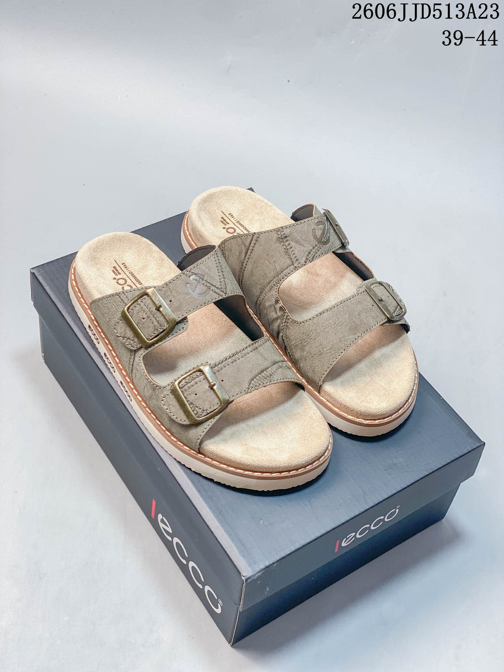 Taupe two-strap slide sandals with buckle details, shown on a shoebox
