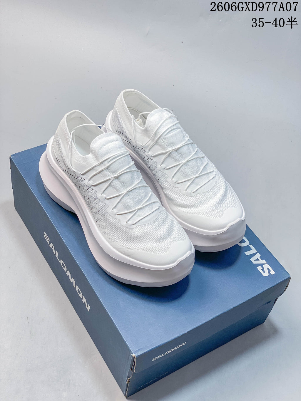 Pair of white Salomon low-top sneakers with cord-style lacing and thick platform soles, displayed on a blue shoebox.