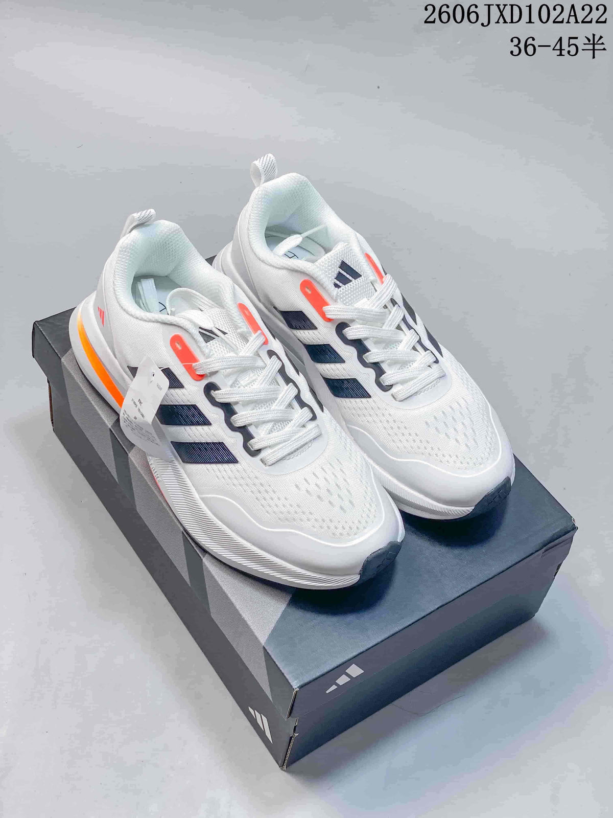 Pair of white Adidas sneakers with dark stripes and orange accents, shown on a shoebox.