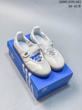 Pair of white Adidas Samba Jane sneakers with pale pink stripes and gum-colored soles, shown on a blue shoe box