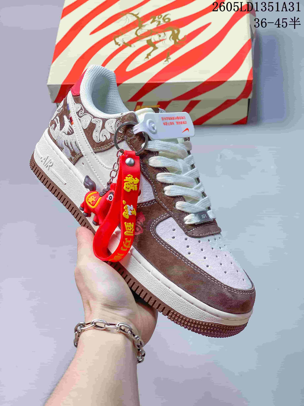 Angled view of the sneaker with embroidered panels, red hanging charm, and patterned box.