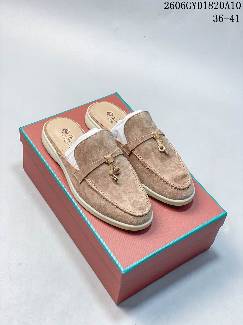Pair of taupe-toned loafer mules with metal vamp details on a coral-colored shoe box