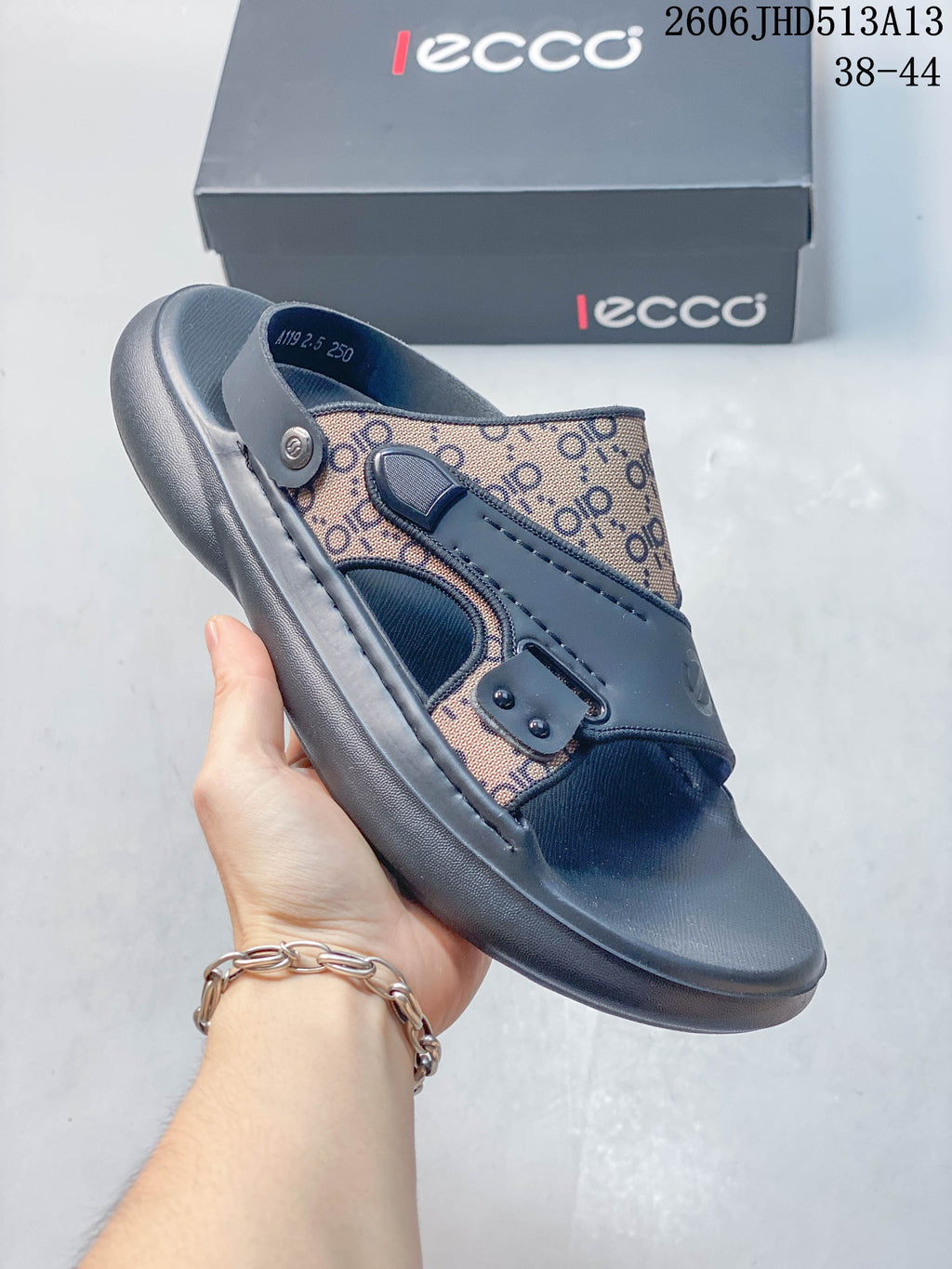 Side view of a gray slide sandal with patterned panels and adjustable-looking strap details, held in front of a branded box.