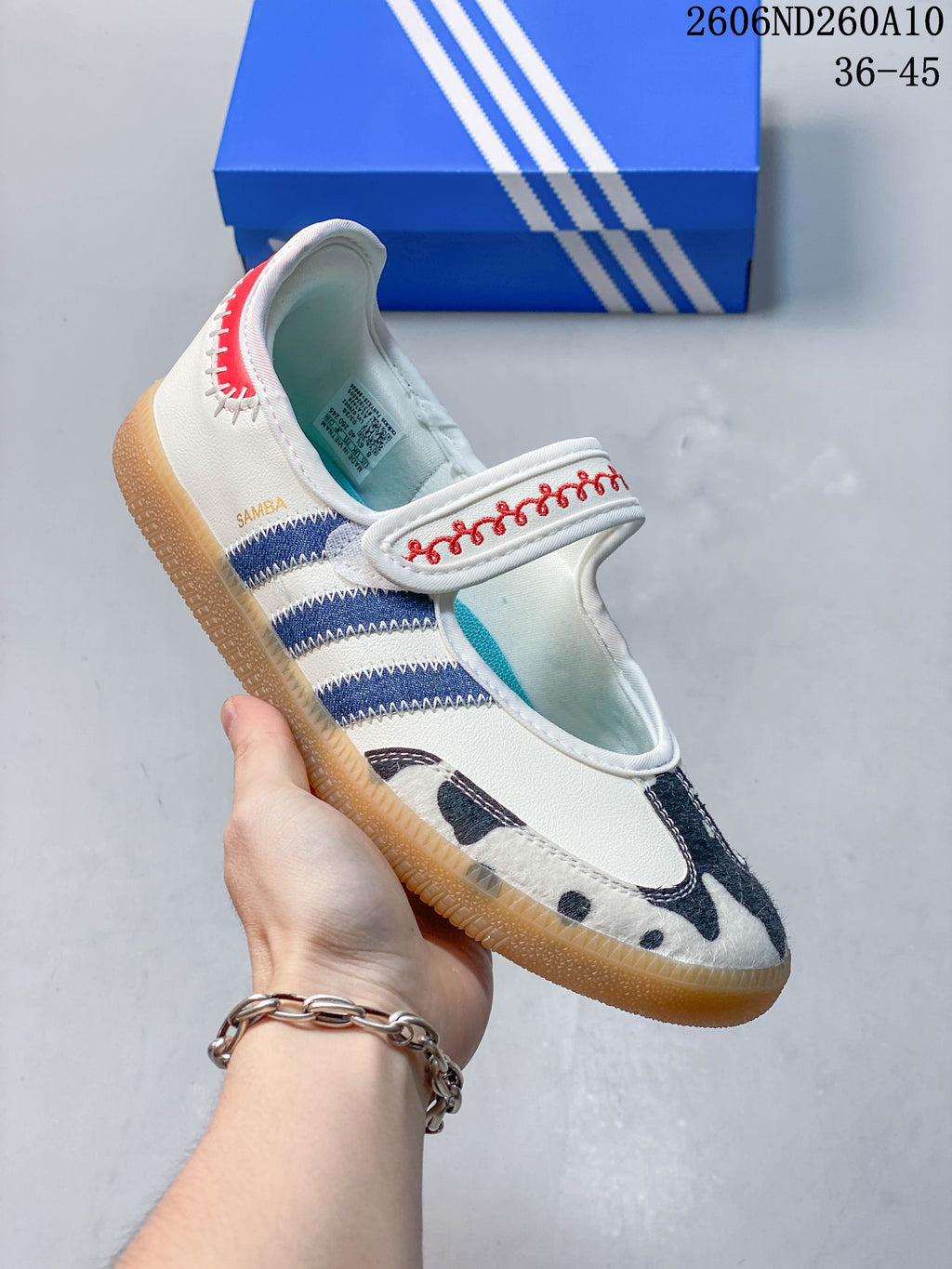 Close-up of an ivory Mary Jane shoe showing a red-embroidered strap, navy side stripes, cow-print toe panel, and gum-colored outsole.