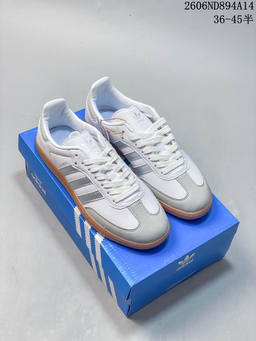 Pair of white adidas Samba OG sneakers with silver details and gum soles, displayed on a blue shoebox