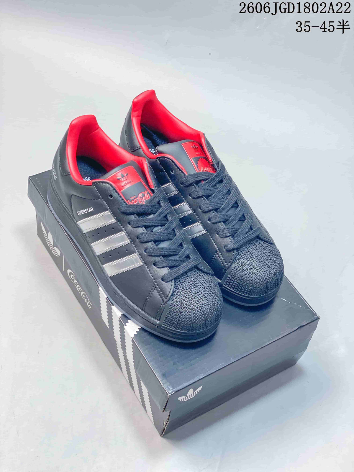Pair of charcoal gray Adidas Originals Superstar sneakers with silver stripes, red accents, and a textured shell toe, shown on a Coca-Cola-branded box.