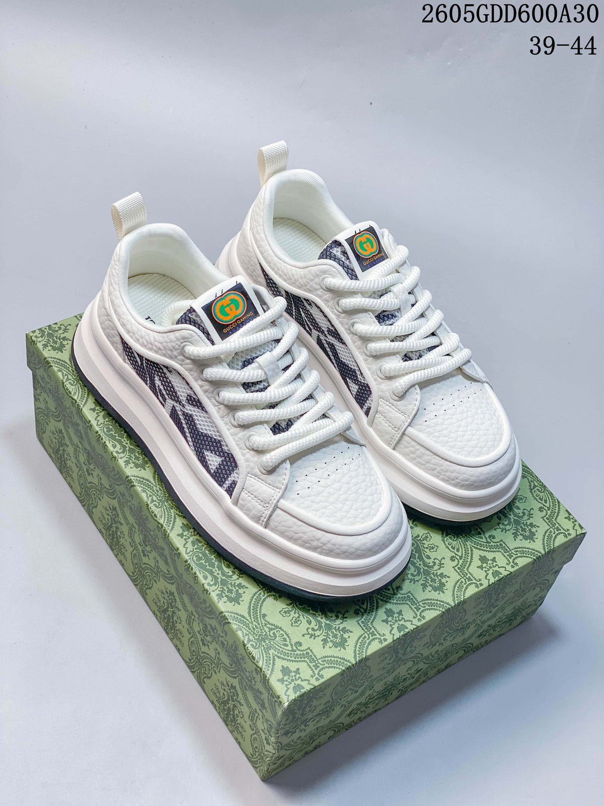 Pair of white Gucci Screener GG-style sneakers with dark patterned side panels, shown on a patterned box.