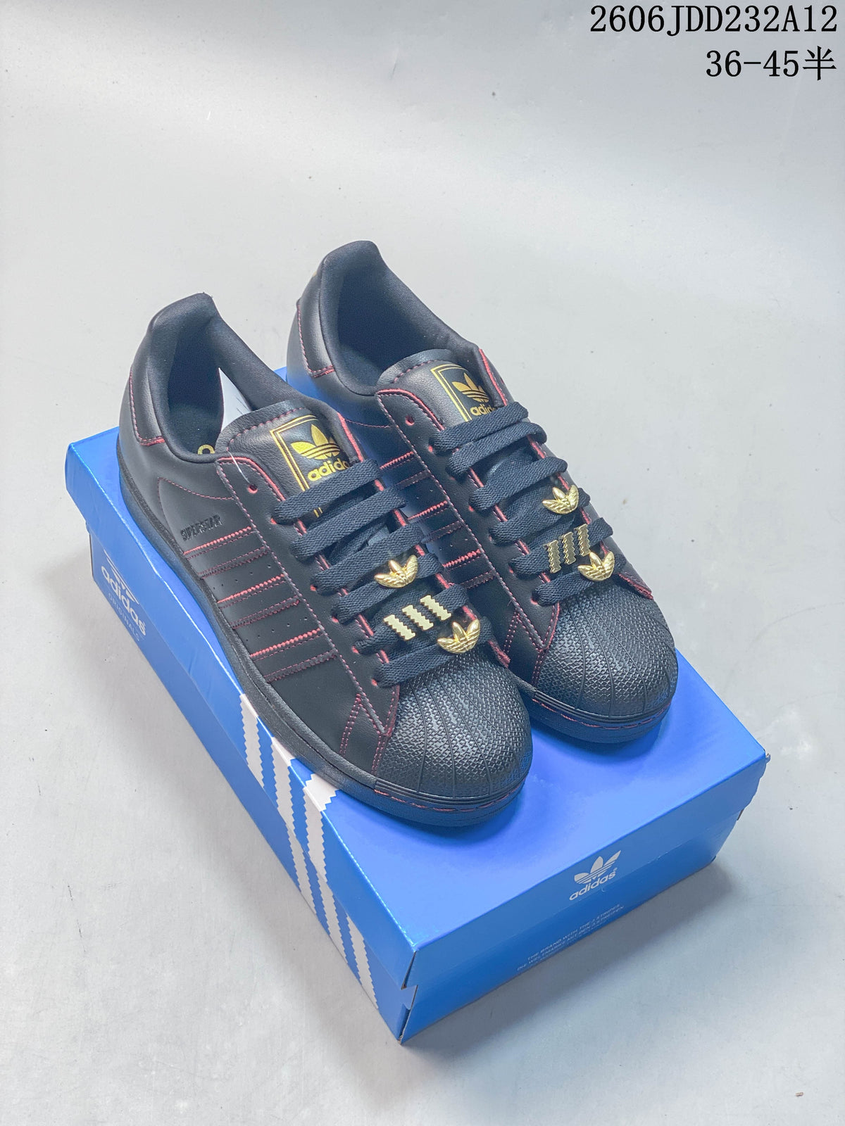 Pair of dark charcoal Adidas Superstar-style low-top sneakers with red stitching and gold-tone lace details, shown on a blue shoe box.