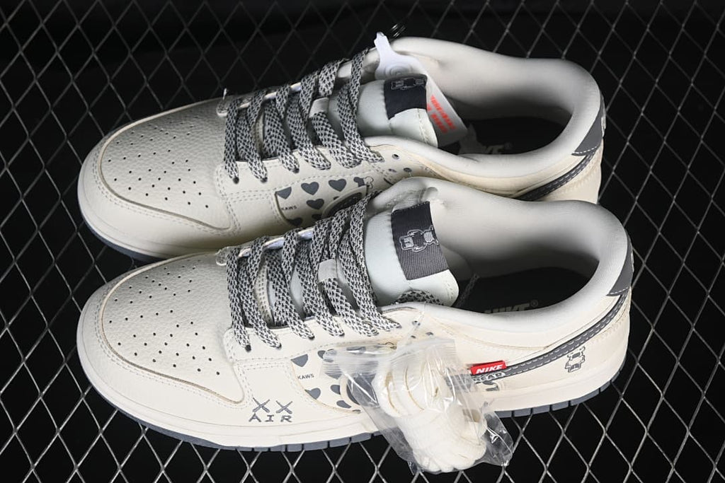 Pair of white and grey Nike SB Dunk Low sneakers with heart-patterned panels, patterned laces, and an additional pair of white laces.