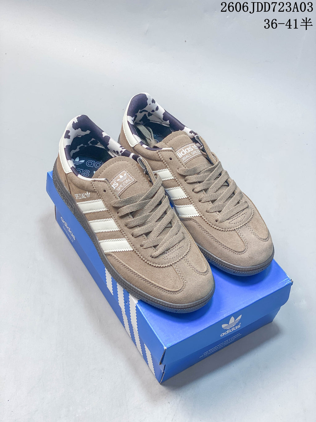Taupe Adidas Handball Spezial sneakers with pale stripes and patterned heel lining, shown on a blue shoe box.