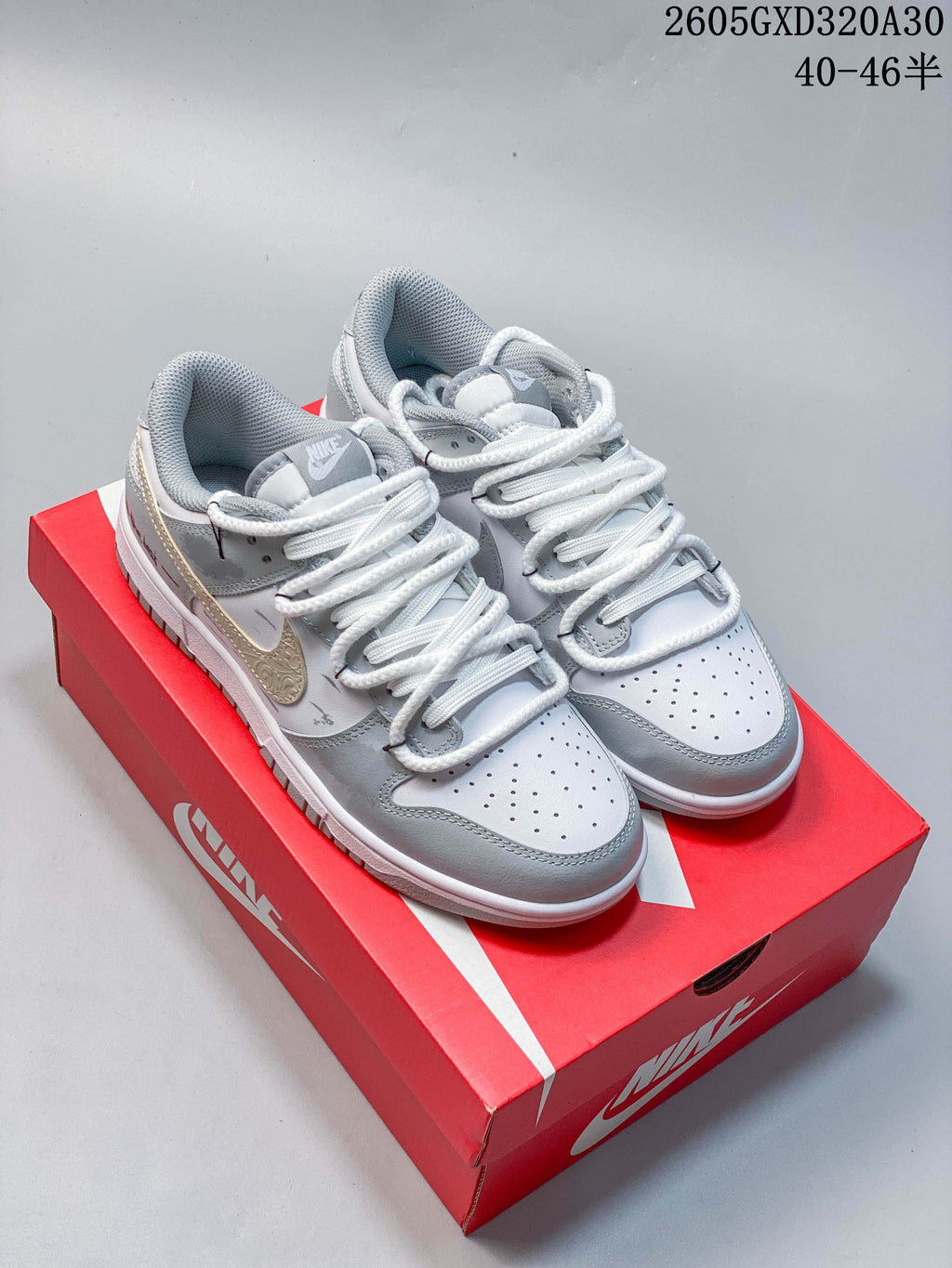 Pair of white and gray low-top Nike Dunk sneakers with a decorative gold-tone Swoosh on a red shoe box.