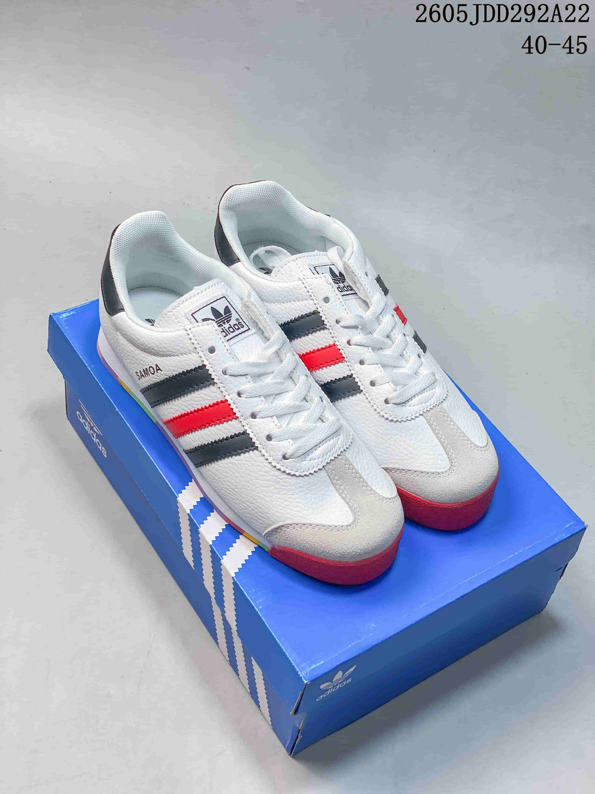 Pair of white Adidas Samoa low-top sneakers with dark gray and red stripes, light toe overlays, and multicolor outsoles on a blue shoe box.