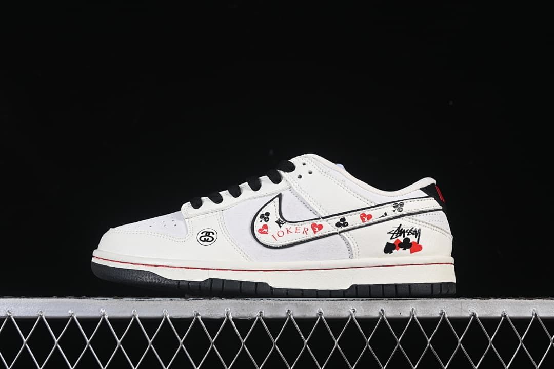 White low-top sneaker with black laces, red accents, playing-card graphics, and Stüssy-style branding, shown in profile.