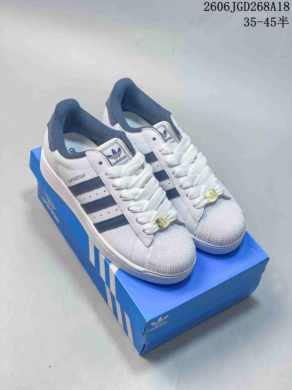 Pair of white Adidas Superstar sneakers with navy stripes and collar lining, shown on a blue shoe box.
