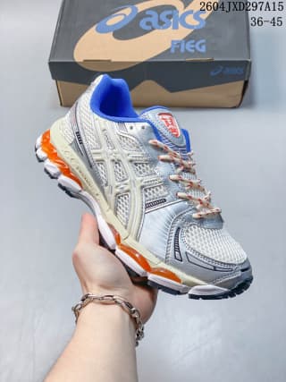 ASICS low-top sneaker held above a branded shoe box, showing the light sage and gray upper, blue collar, orange midsole accents, and style marking 1203B144-102.
