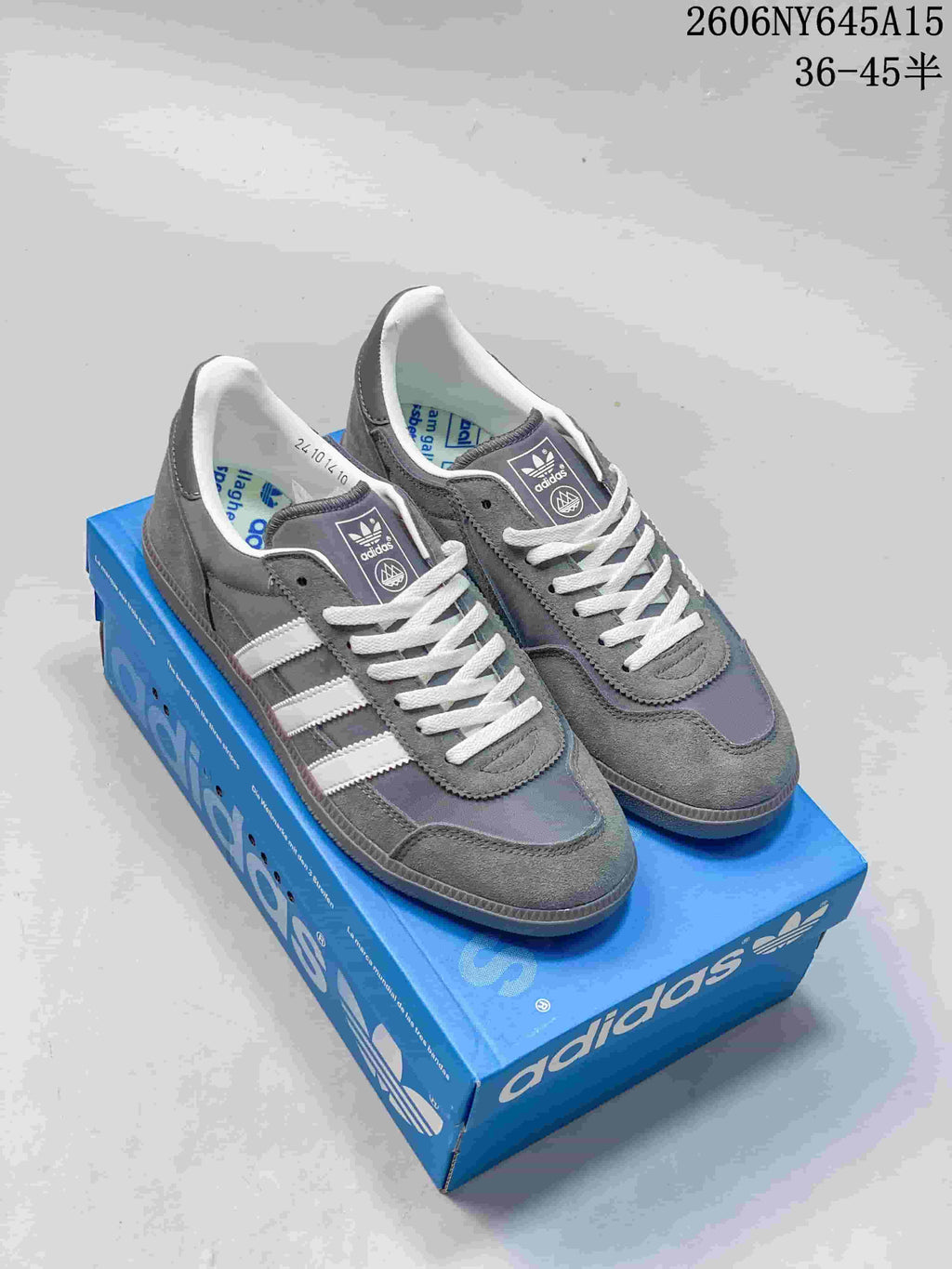 Pair of grey adidas Handball Spezial style sneakers with white stripes and laces on a blue shoebox