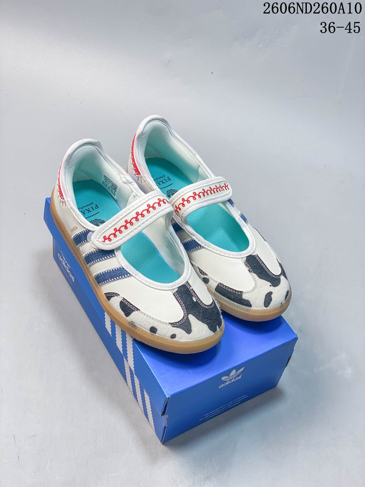 Ivory adidas Samba Jane-style Mary Jane shoes with turquoise insoles, red strap embroidery, navy stripes, and cow-print toe panels on a blue shoe box.