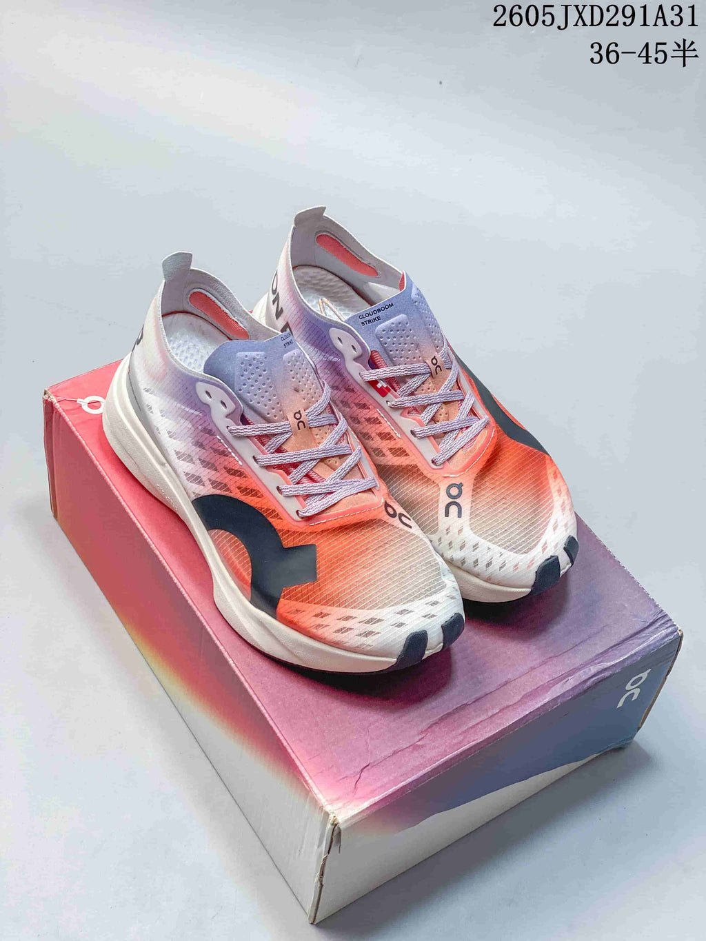 Pair of On Cloudboom Strike running shoes in coral, lilac, white, and dark gray, shown on a shoebox.